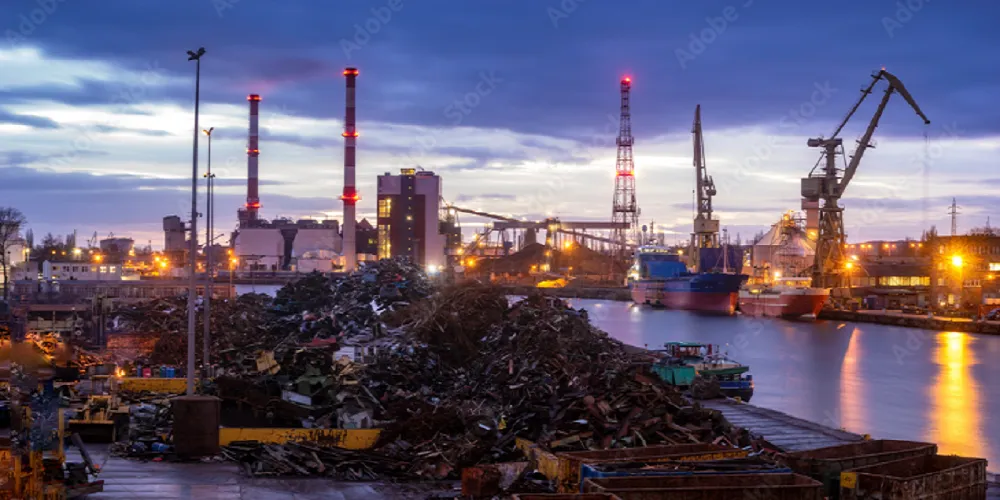 ميناء صناعي ليلاً تظهر فيه أكوام ضخمة من الخردة المعدنية بجوار سفن شحن ورافعات عملاقة ومداخن مصانع مضاءة، في مشهد يعبر عن تجارة سكراب الحديد والنحاس والألمنيوم ونقلها عبر البحر لإعادة التدوير، مما يبرز نشاط شراء سكراب الطائف ودور الموانئ في دعم الاقتصاد والصناعات الثقيلة وحماية البيئة من النفايات المعدنية وتحقيق الاستدامة